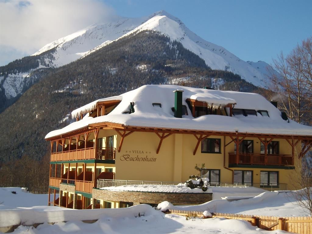 A charming hotel with a snow-covered roof and cozy balconies. In the background, majestic mountains can be seen under a blue sky.