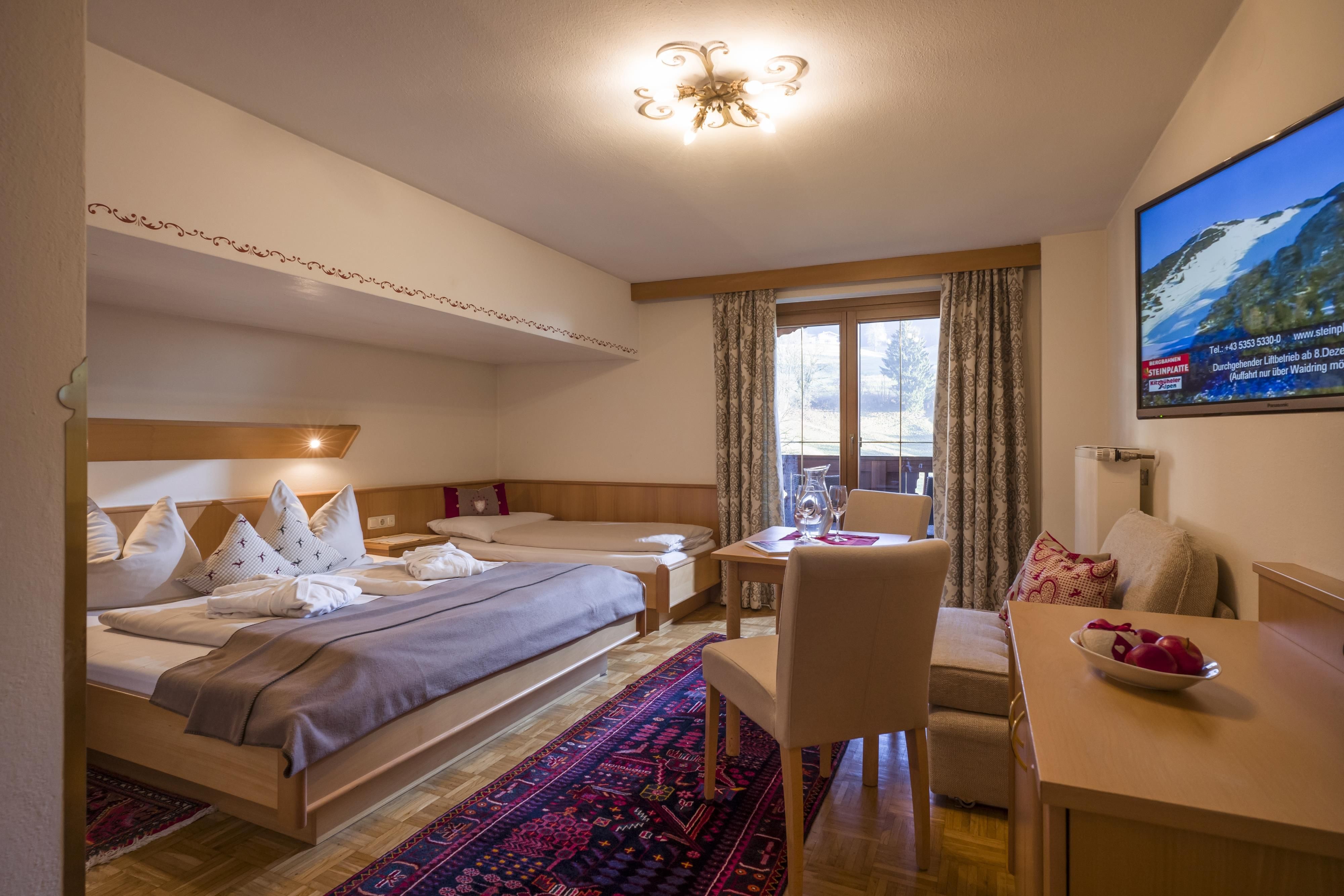 A cozy bedroom with two beds and a small table. The room has a balcony and a television on the wall.