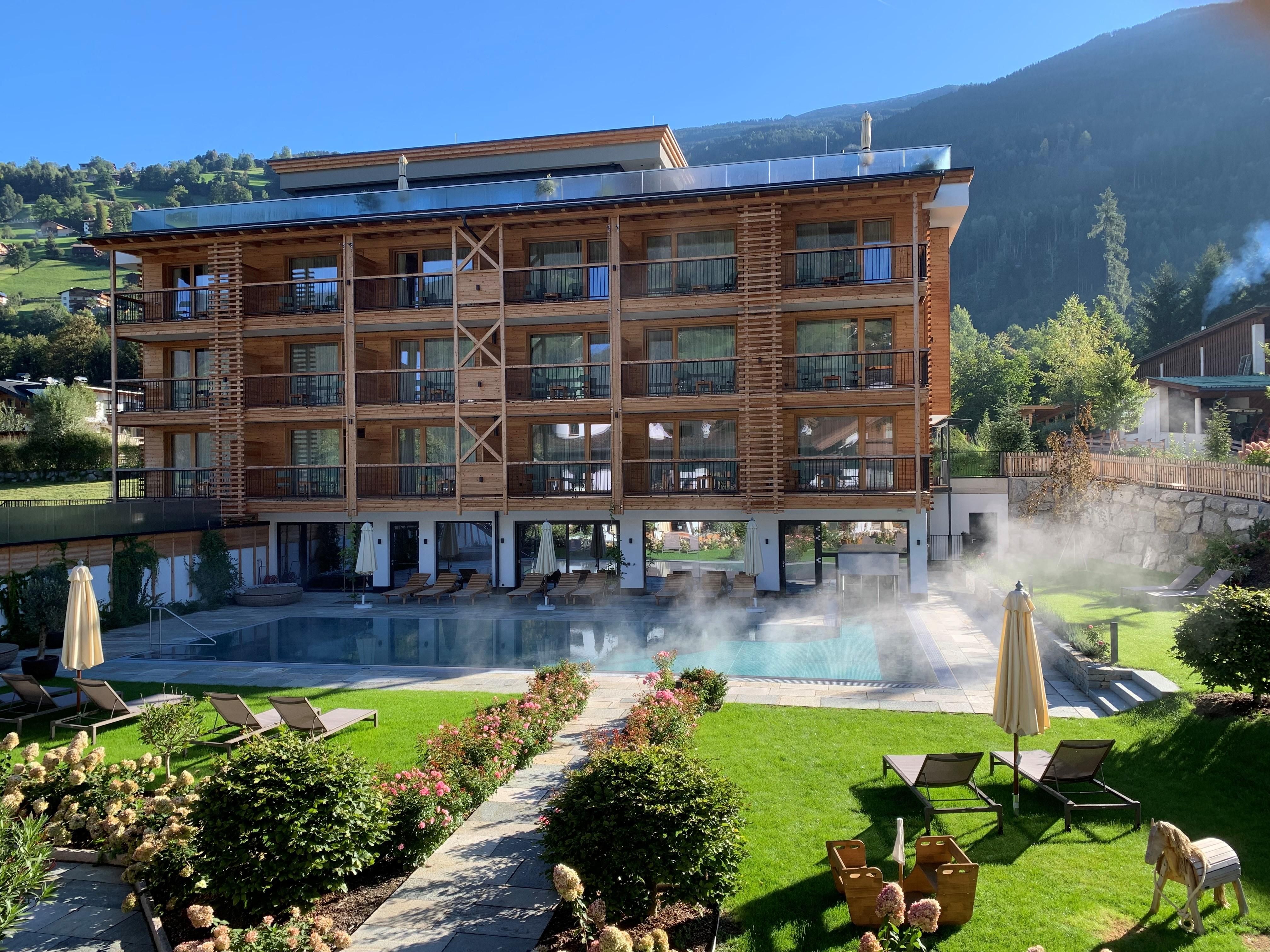 A modern hotel with a wooden facade and large windows. In front of the hotel, there is a pool and a well-maintained garden landscape.