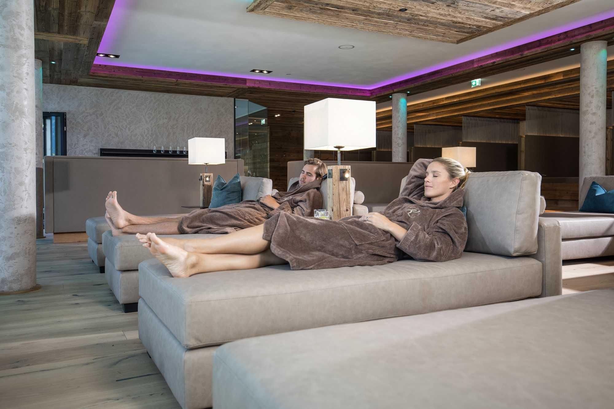 Two people relax in a modern wellness area. They are wearing bathrobes and lying on comfortable lounge furniture.