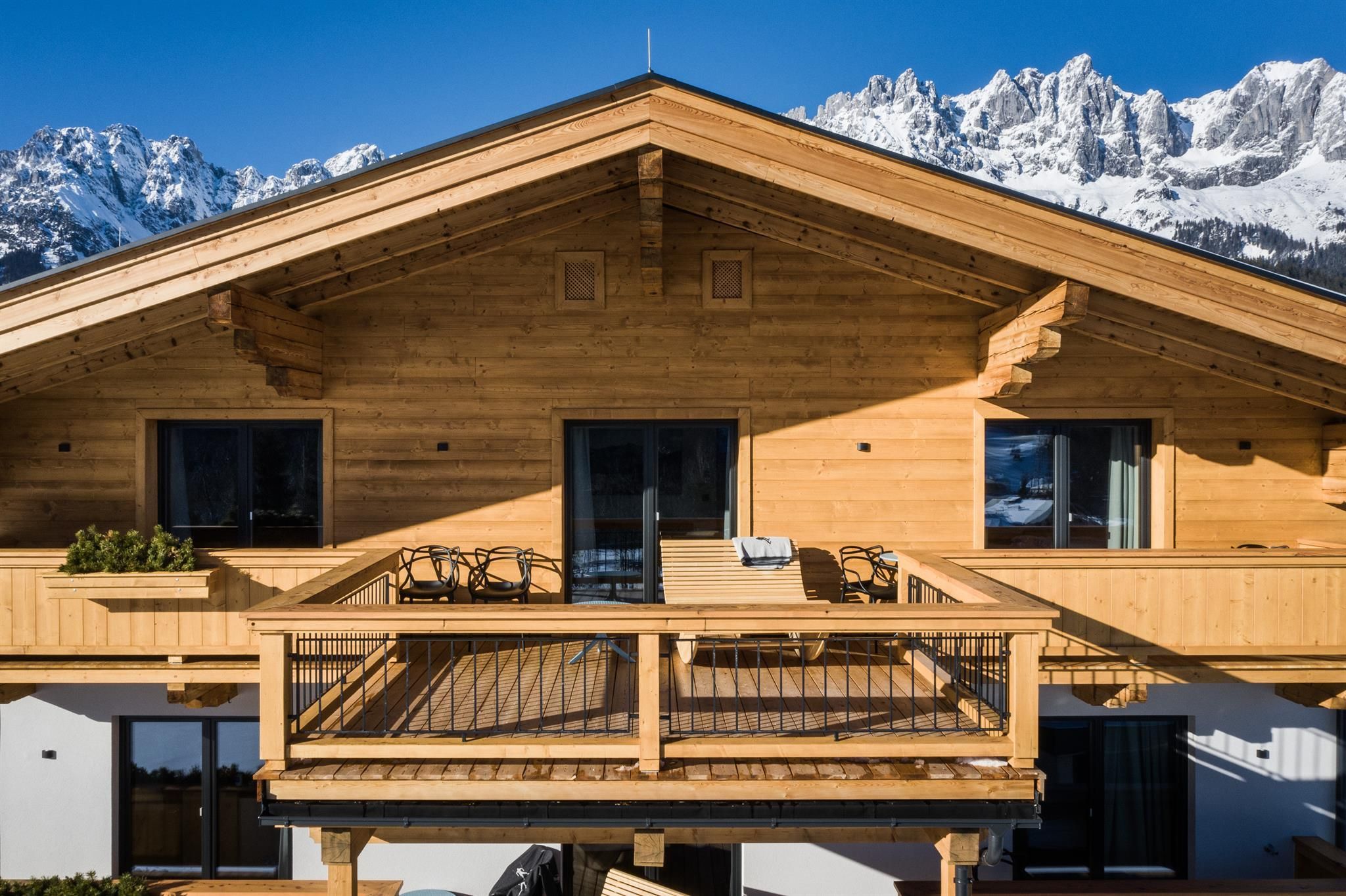A modern wooden house with a large balcony and a view of the snow-covered mountains. The facade is bright and inviting.