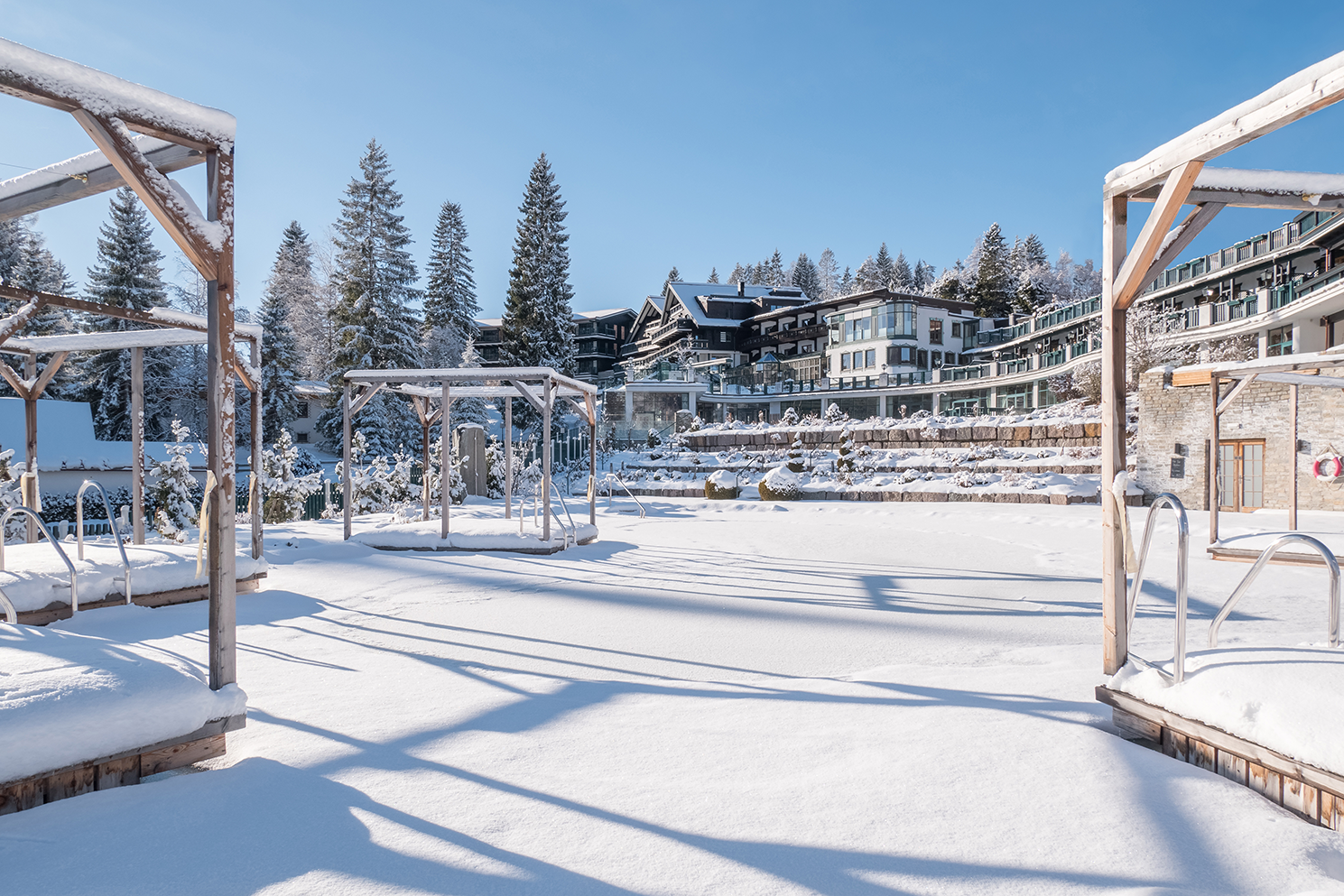 A peaceful conservatory with a snow-covered area and clear blue sky. In the background, modern buildings and snow-covered trees are visible.