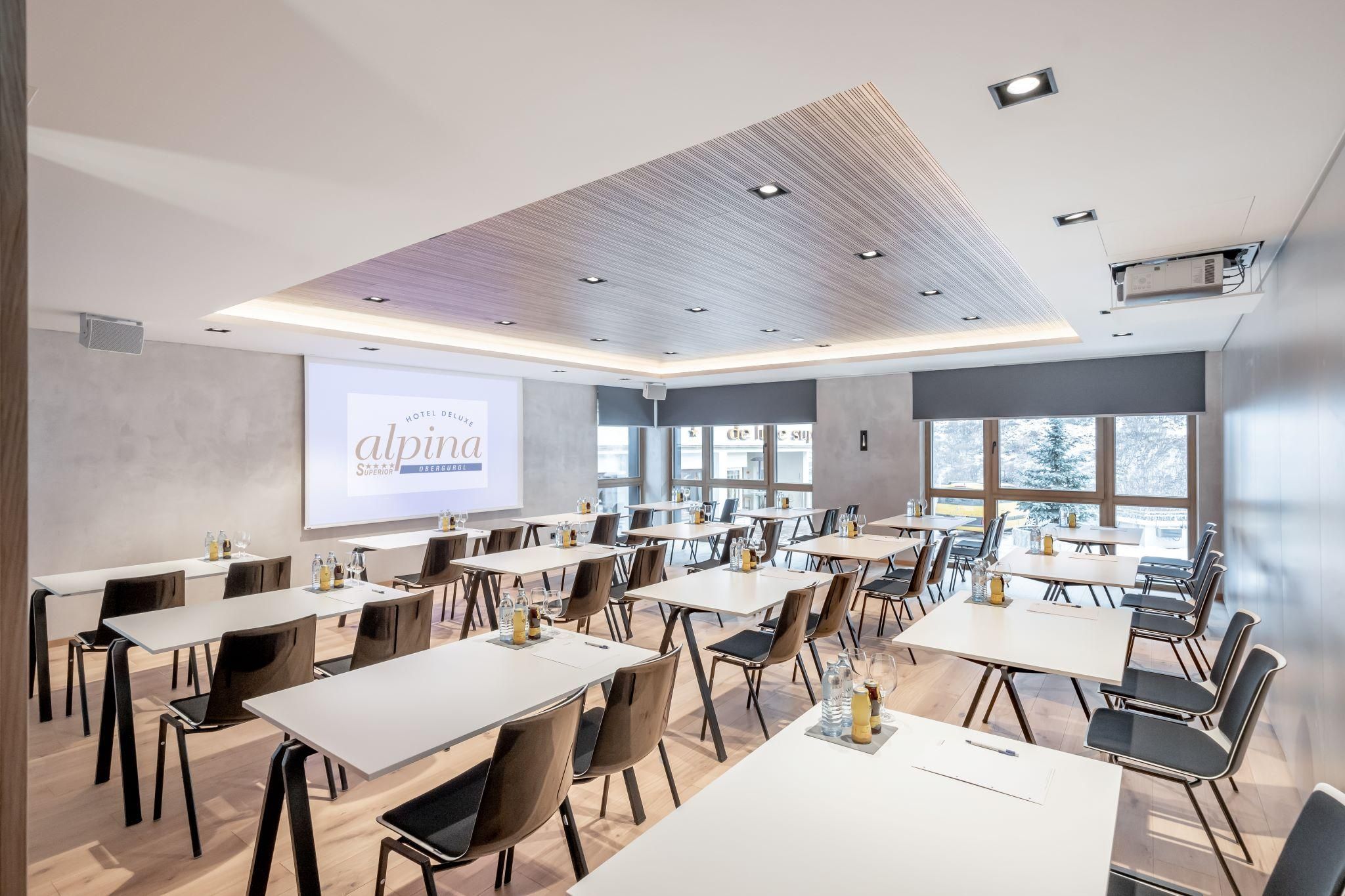 A modern conference room with bright walls and large windows. There are several tables and chairs, as well as a projector for presentations.