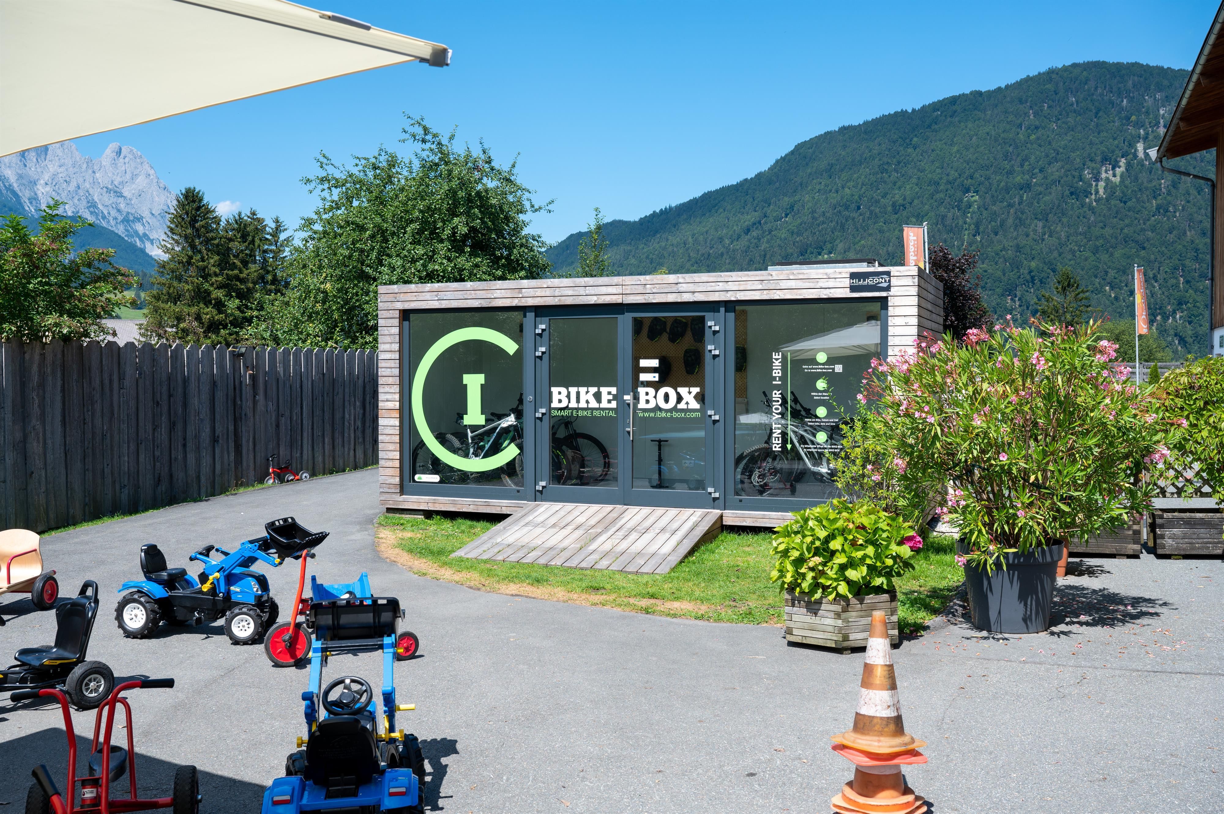 A modern bike shop called "Bike Box" in a picturesque setting with mountains in the background. In front of the store, there are several children's bikes and blooming plants.