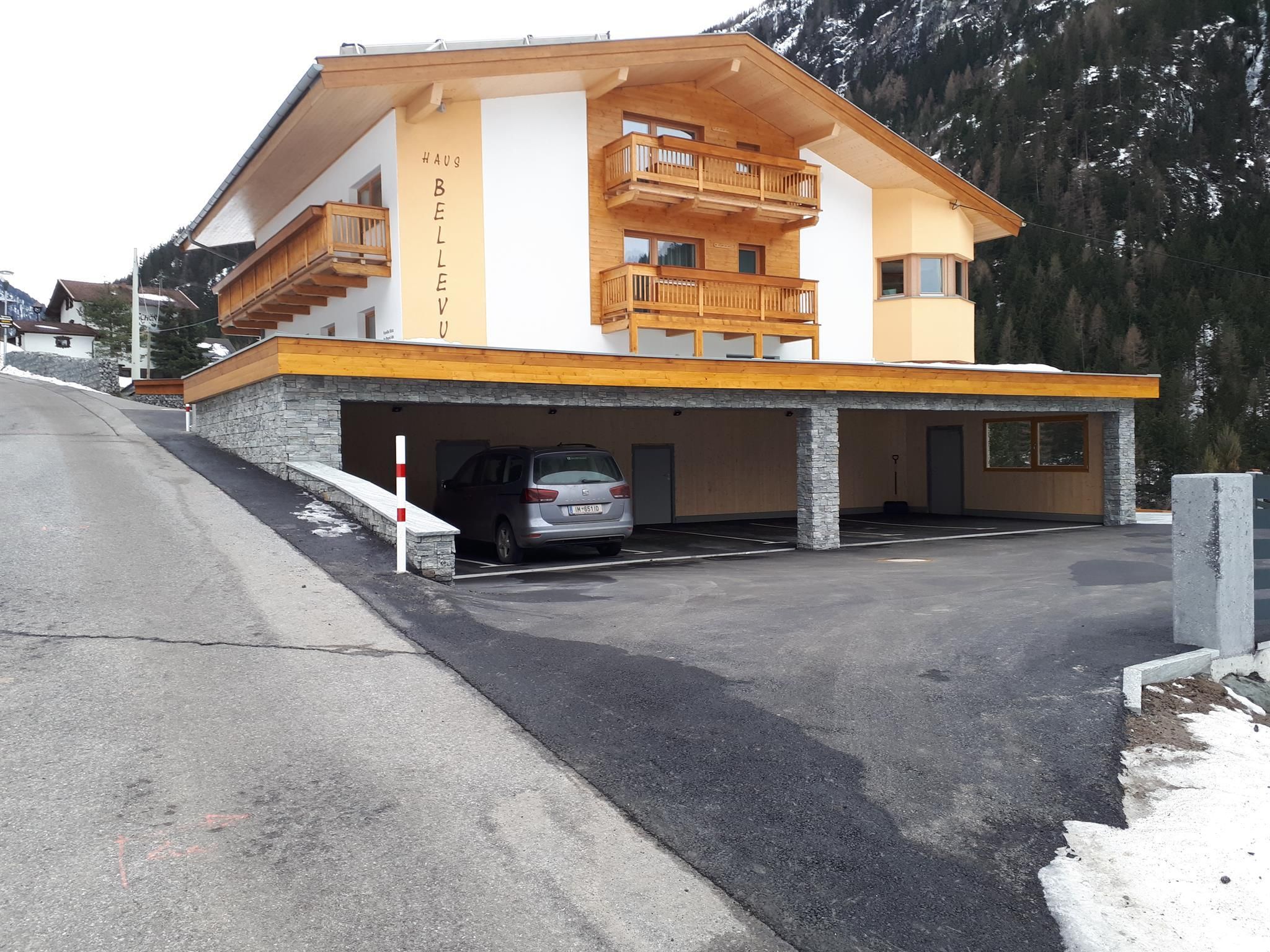 A modern building in the mountains with a covered parking area. There are several balconies and a striking wood cladding.