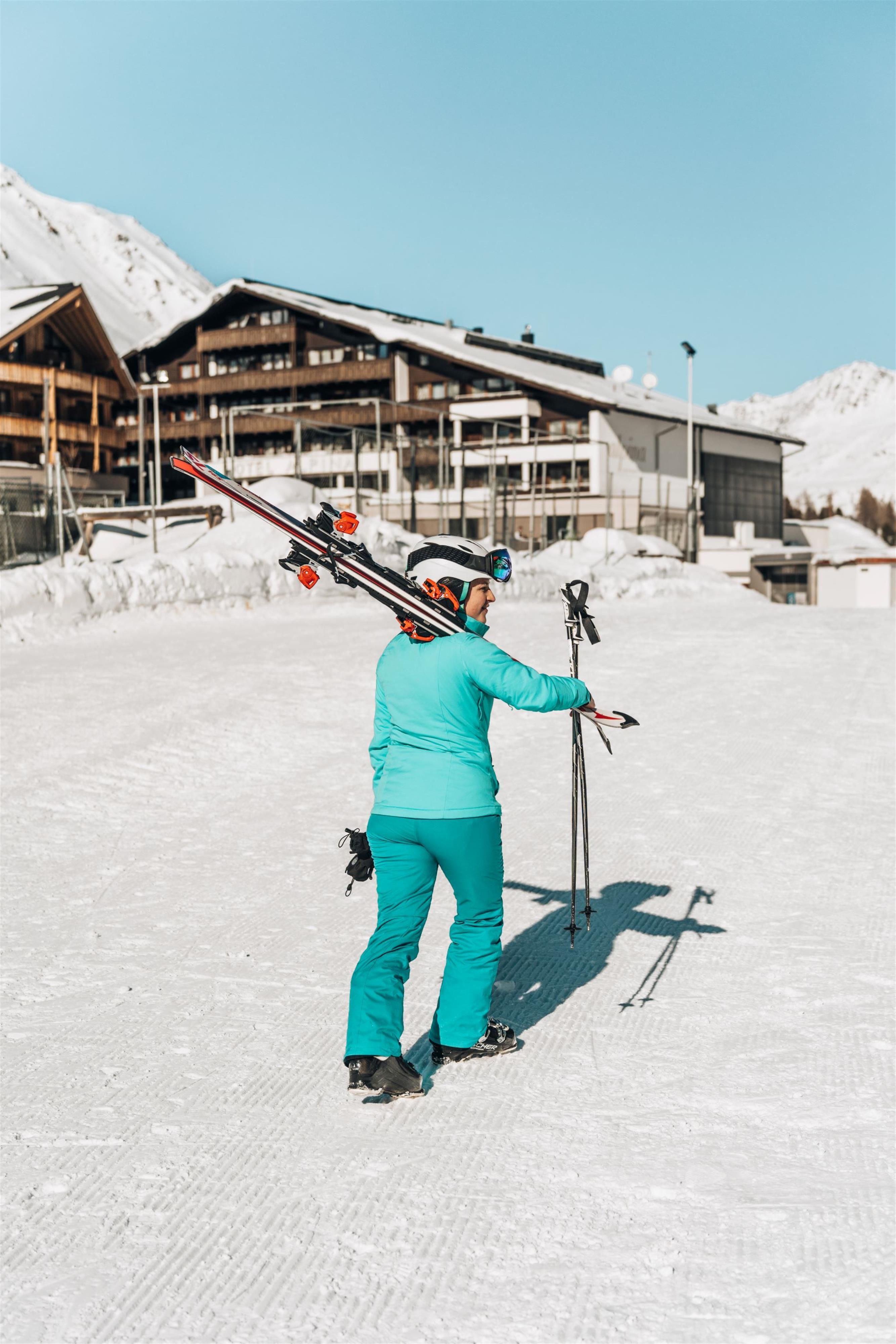 A person in blue-green ski clothing is carrying skis on their shoulder. In the background, snow-covered mountains and ski huts can be seen.