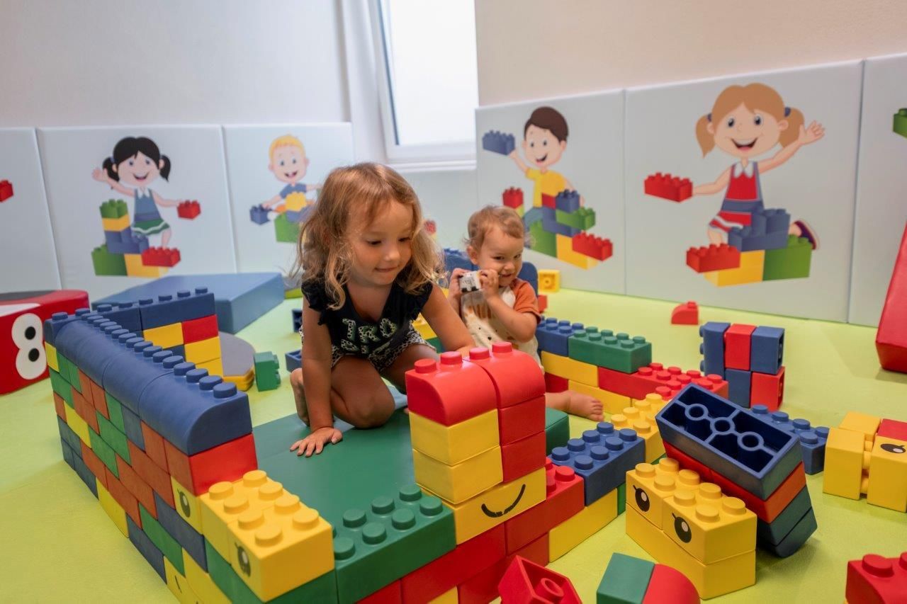 Two small children are playing with colorful building blocks in a playroom. In the background, cheerful wall murals of other children can be seen.