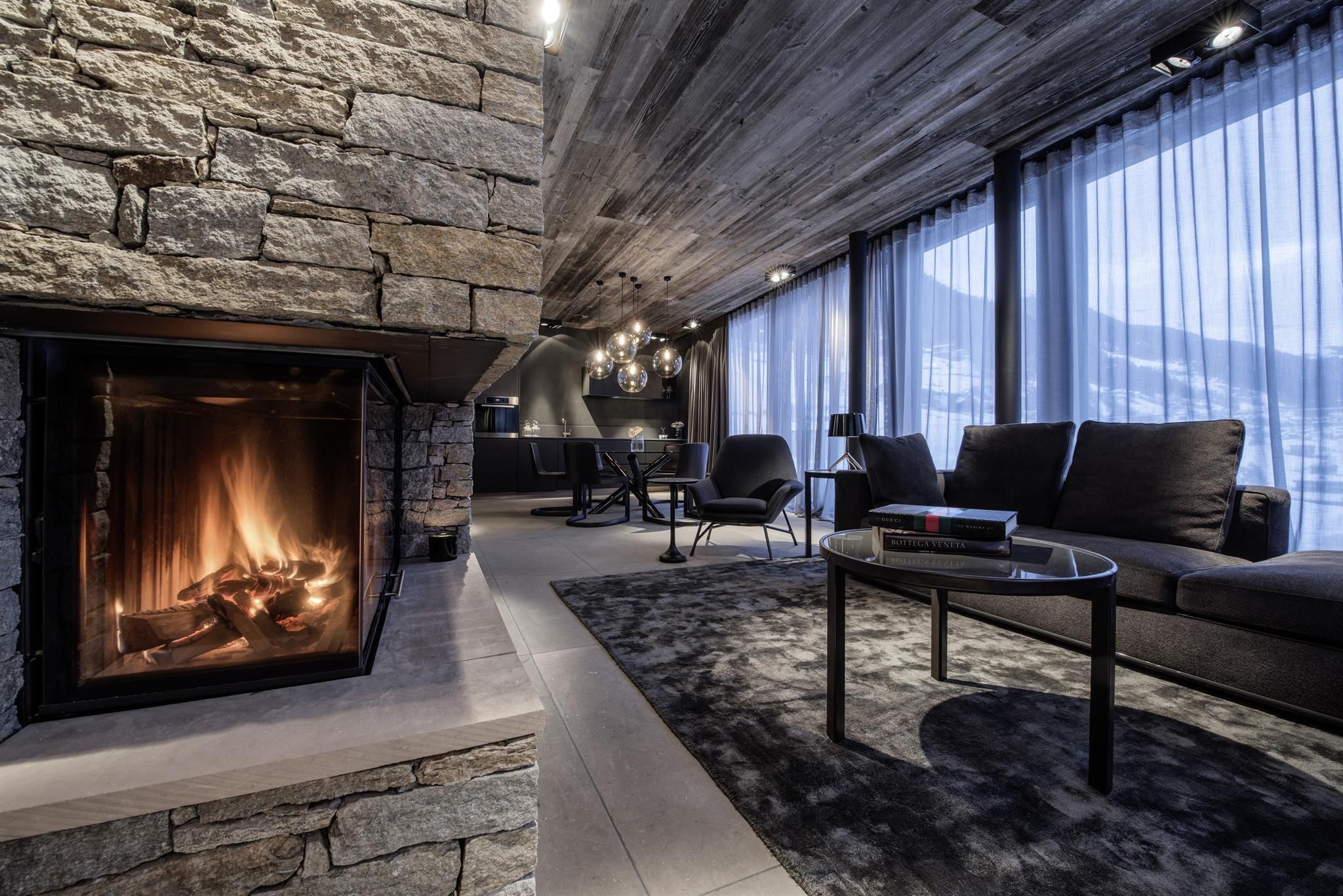 A modern living room with an elegant stone fireplace and large windows. The dark furniture and the carpet create a cozy atmosphere.