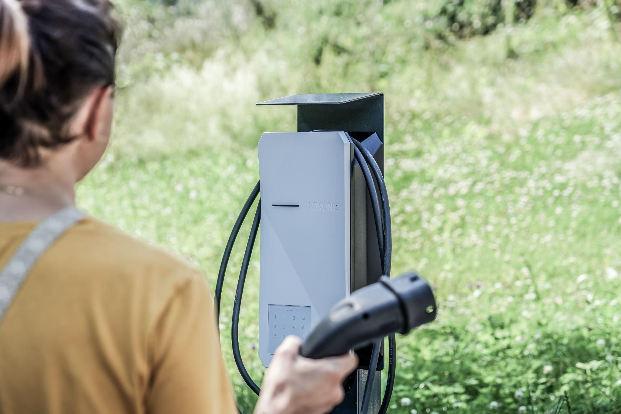 A person stands in front of a charging station for electric vehicles. In the background, green meadows and trees can be seen.