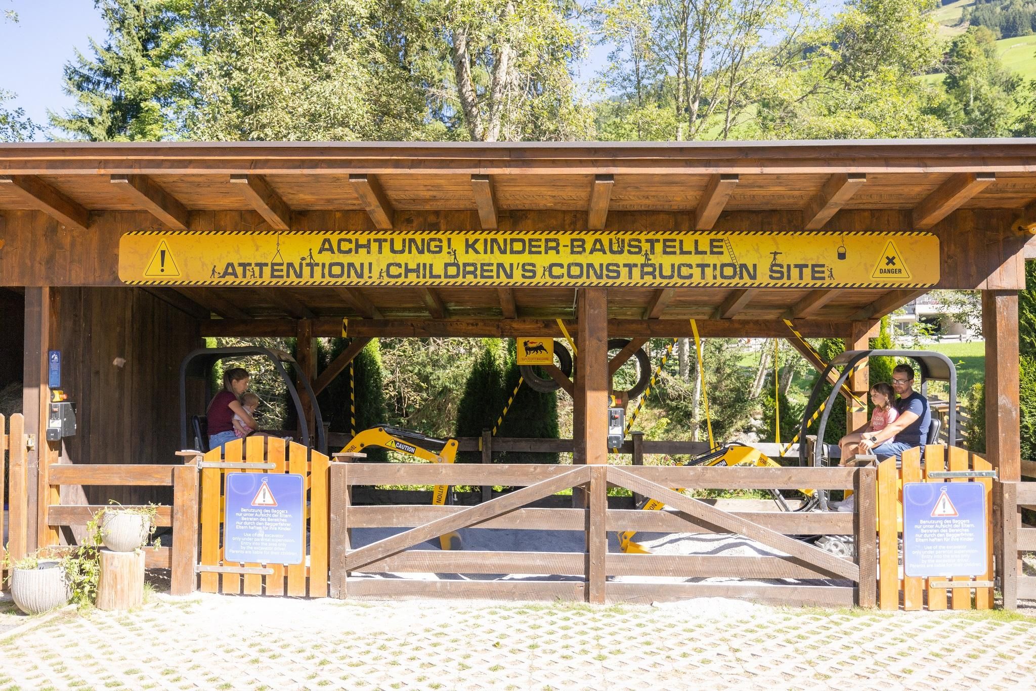 An entrance to a children's construction site with a large yellow sign. In the background, play equipment and a green environment are visible.