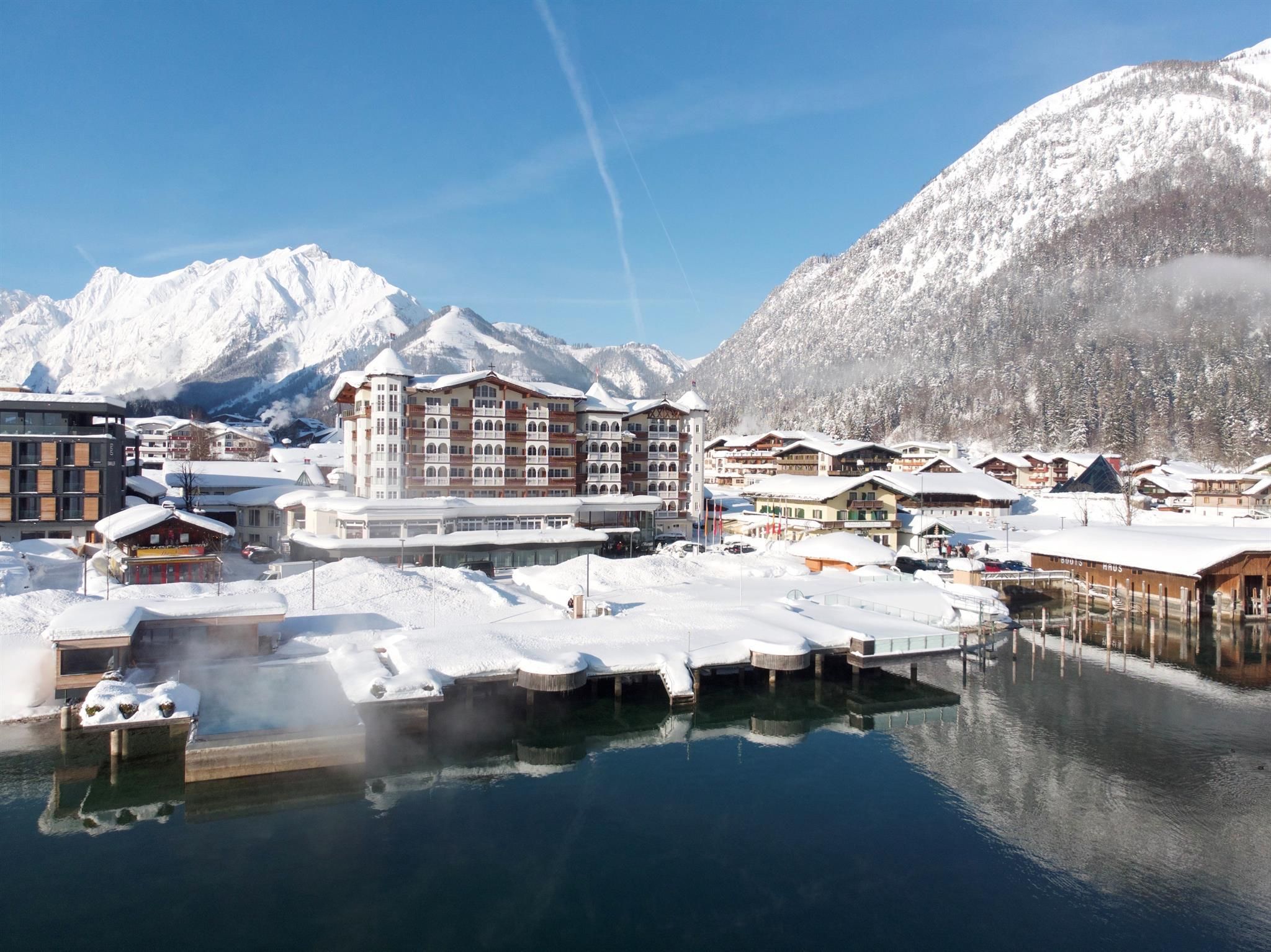 An idyllic winter landscape with snow-covered mountains and a tranquil lake. In the foreground, modern buildings and boat docks stand, surrounded by fresh, white snow.