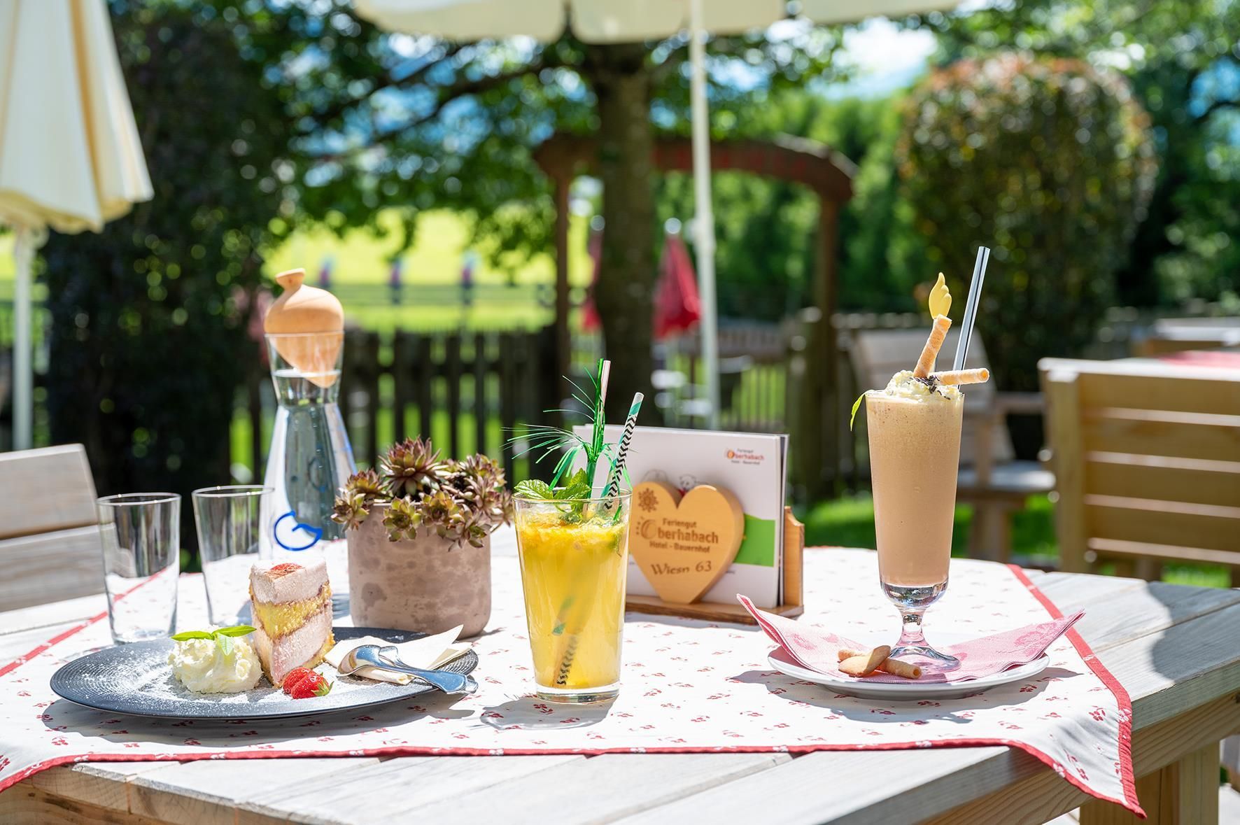 A lovingly set outdoor table with two delicious drinks and a dessert. In the background, green meadows and trees can be seen.