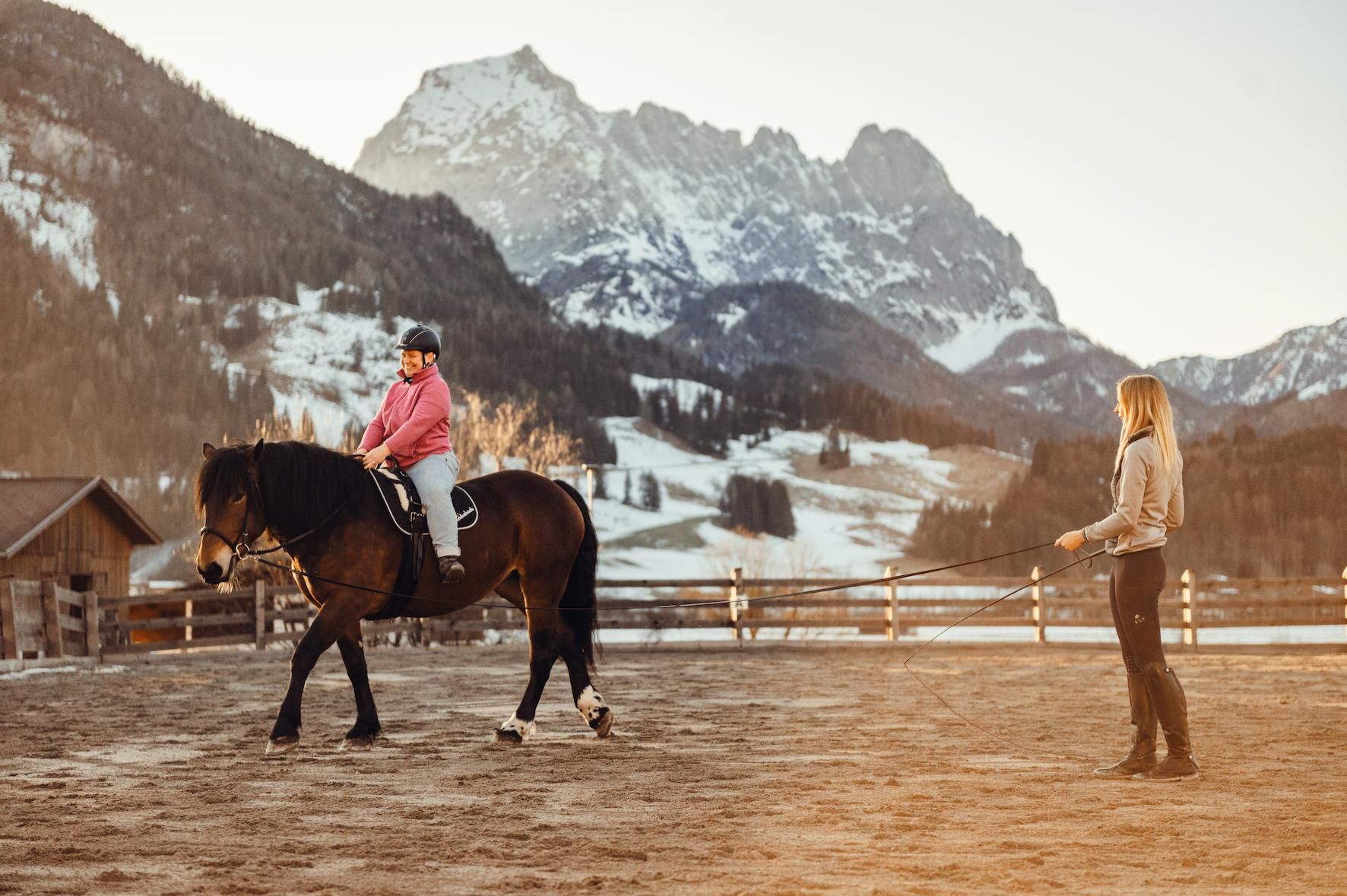 A child rides on a horse in a riding hall with picturesque mountains in the background. A woman stands next to the horse and watches the rider.