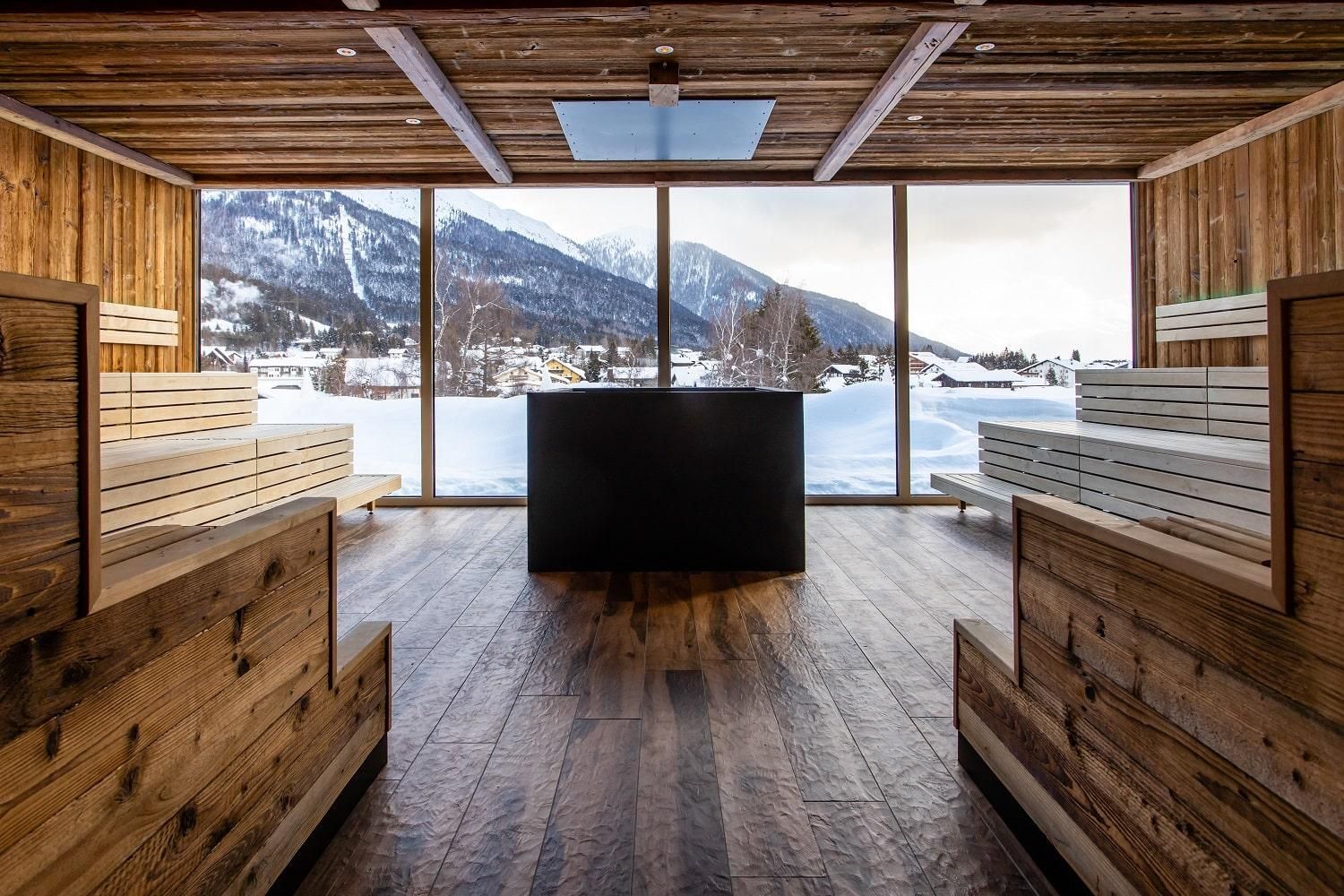 A modern, wood-paneled room with panoramic windows overlooking snow-covered mountains. In the center stands a black piece of furniture, surrounded by wooden benches.