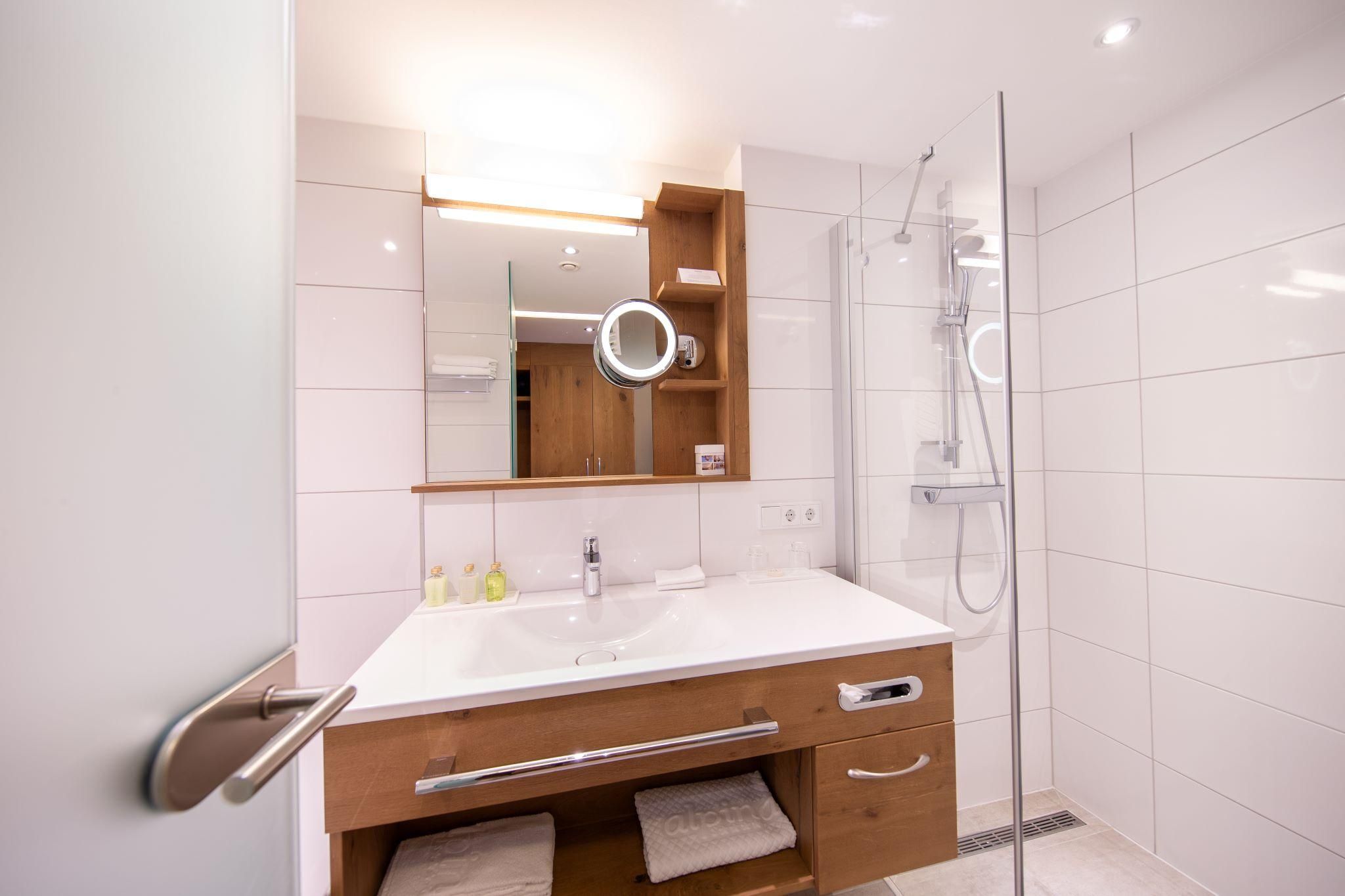 A modern bathroom with a bright shower and a large mirror. The furniture is made of wood and the walls are light tiled.