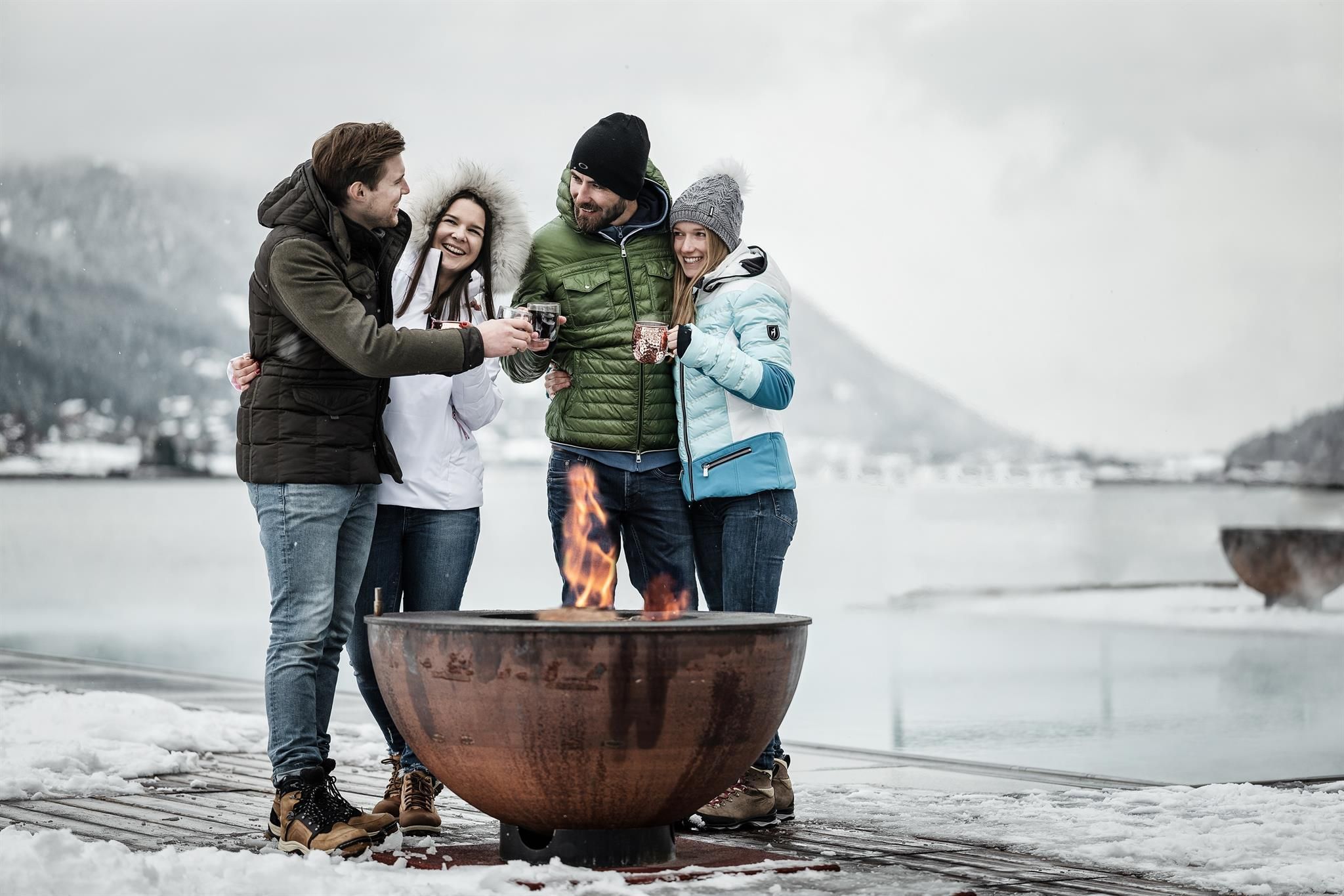 A group of four friends stands around a fire in the snow. They laugh and enjoy the frosty surroundings with mountains in the background.