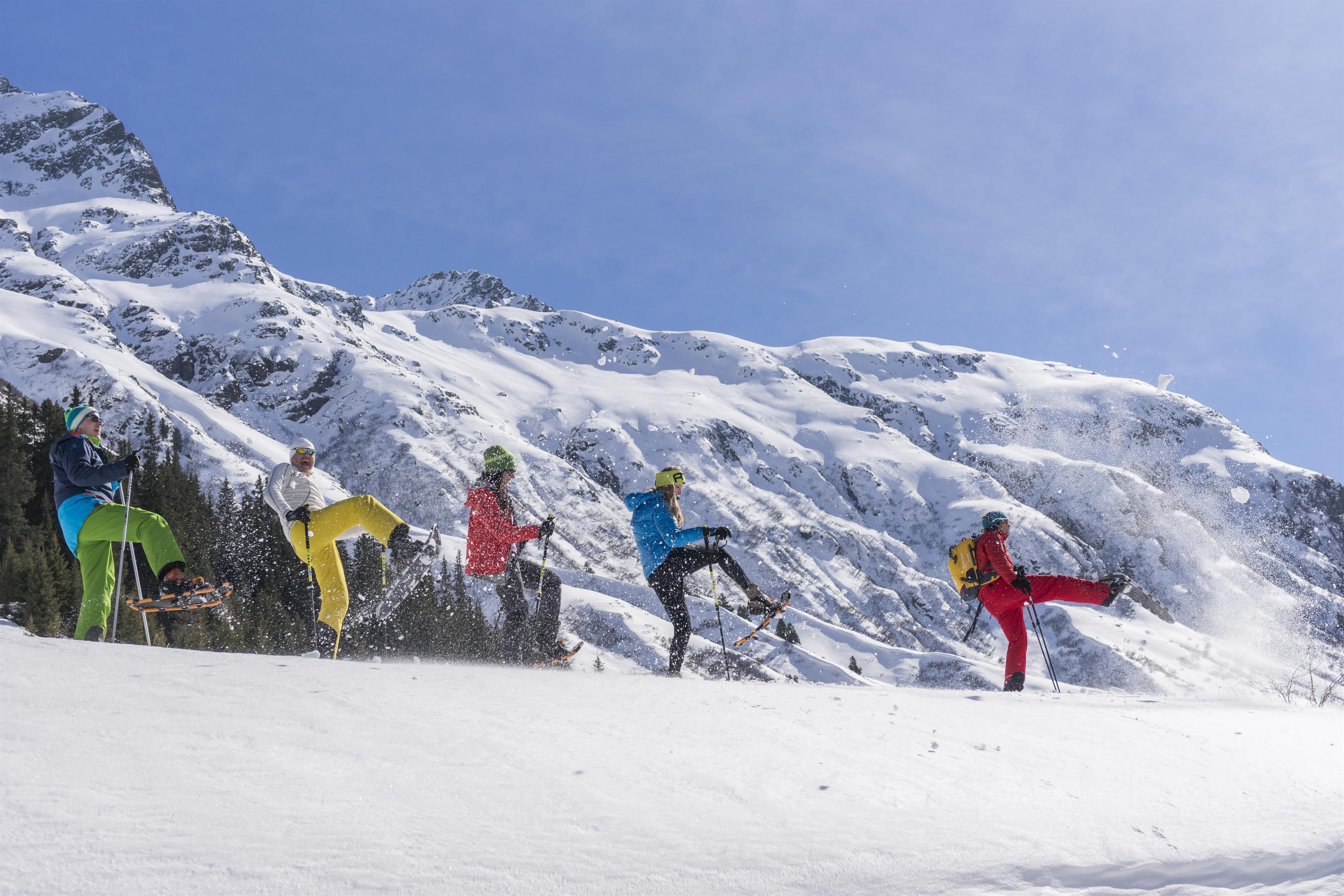 A group of skiers in colorful outfits is enjoying a sunny day in the snow. Snow-covered mountains can be seen in the background.