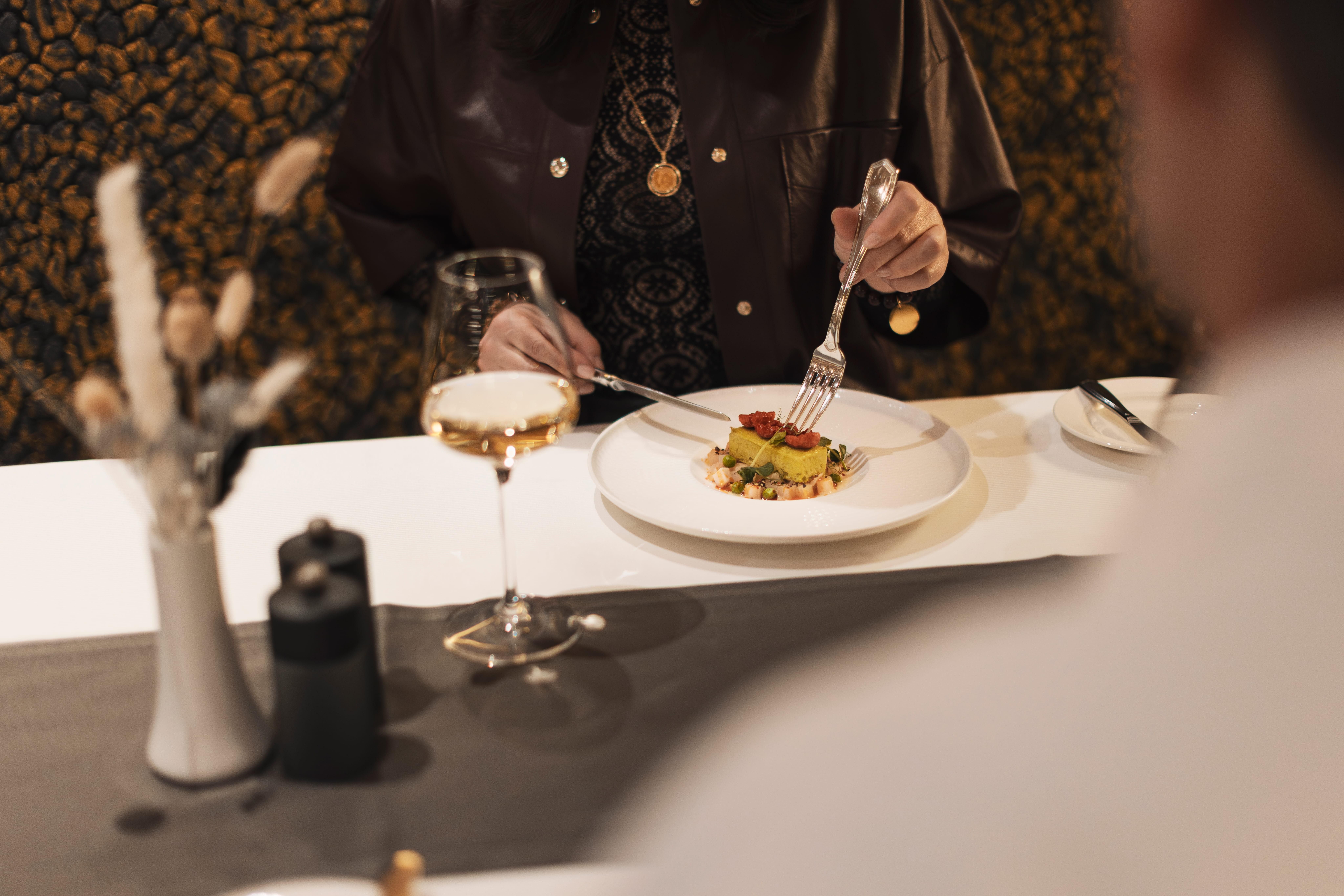 A person is sitting at a table with an elegant dish. They are holding cutlery and enjoying a glass of wine.