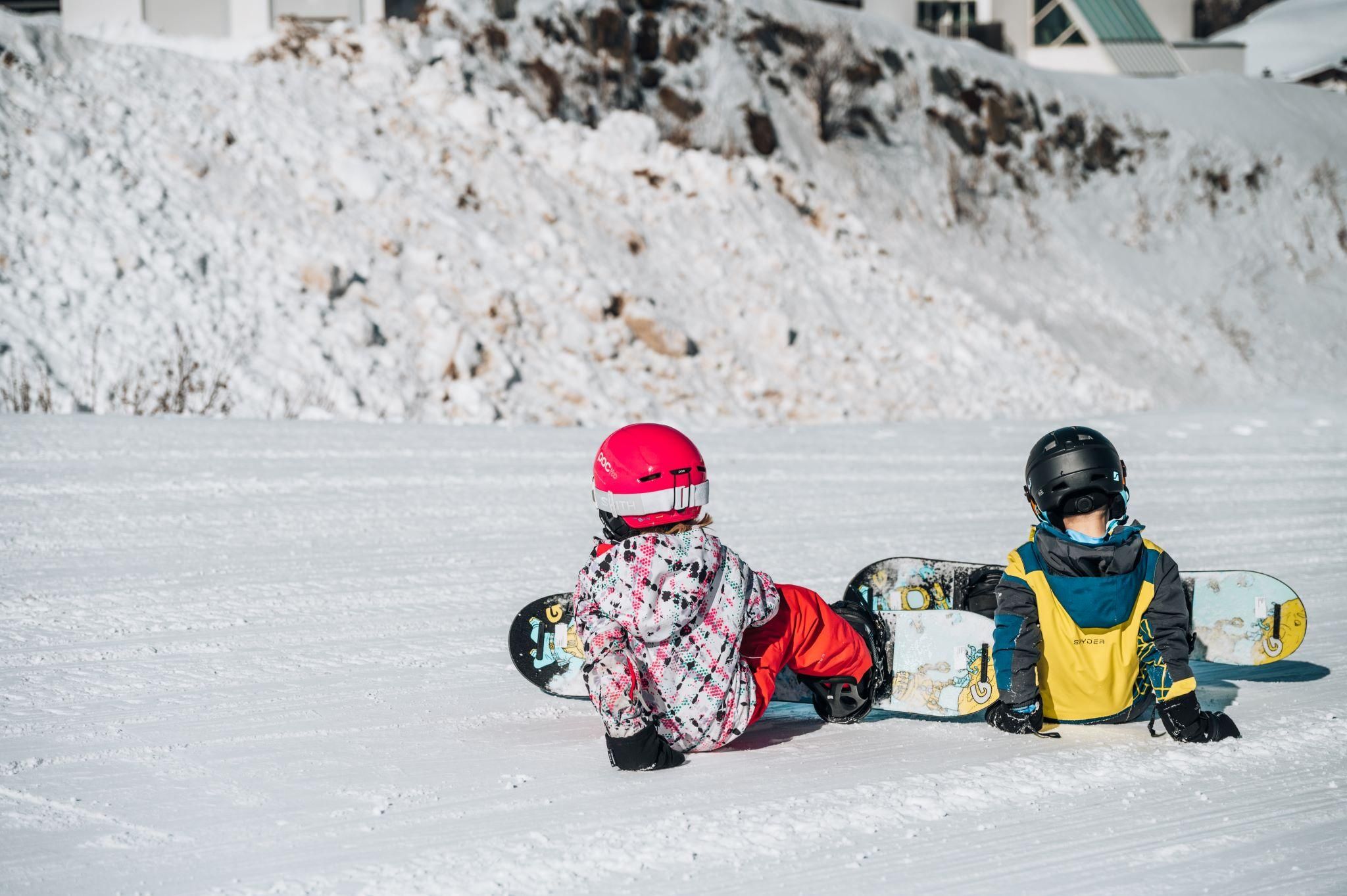 Two children are sitting in the snow holding snowboards. They are wearing colorful winter clothing and helmets.