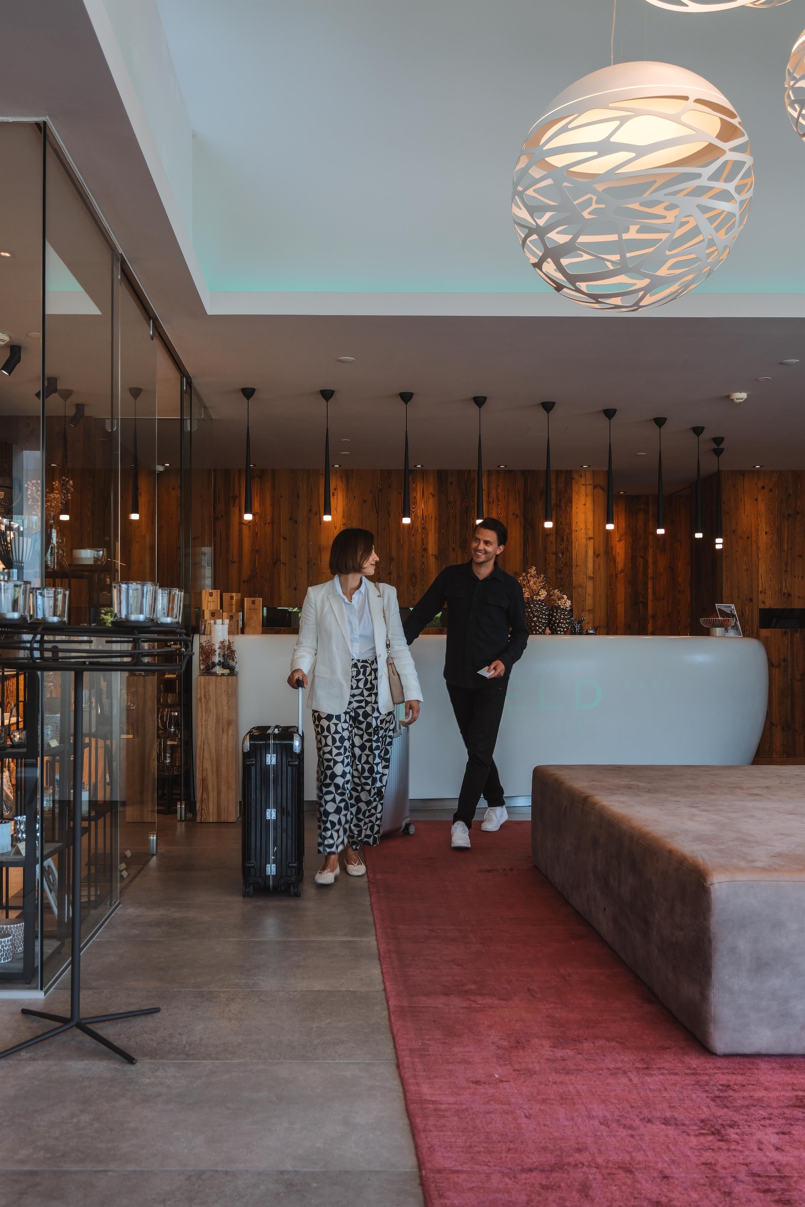 A modern hotel lobby with a reception desk. A man and a woman are standing at the reception, while the woman arrives with her luggage.