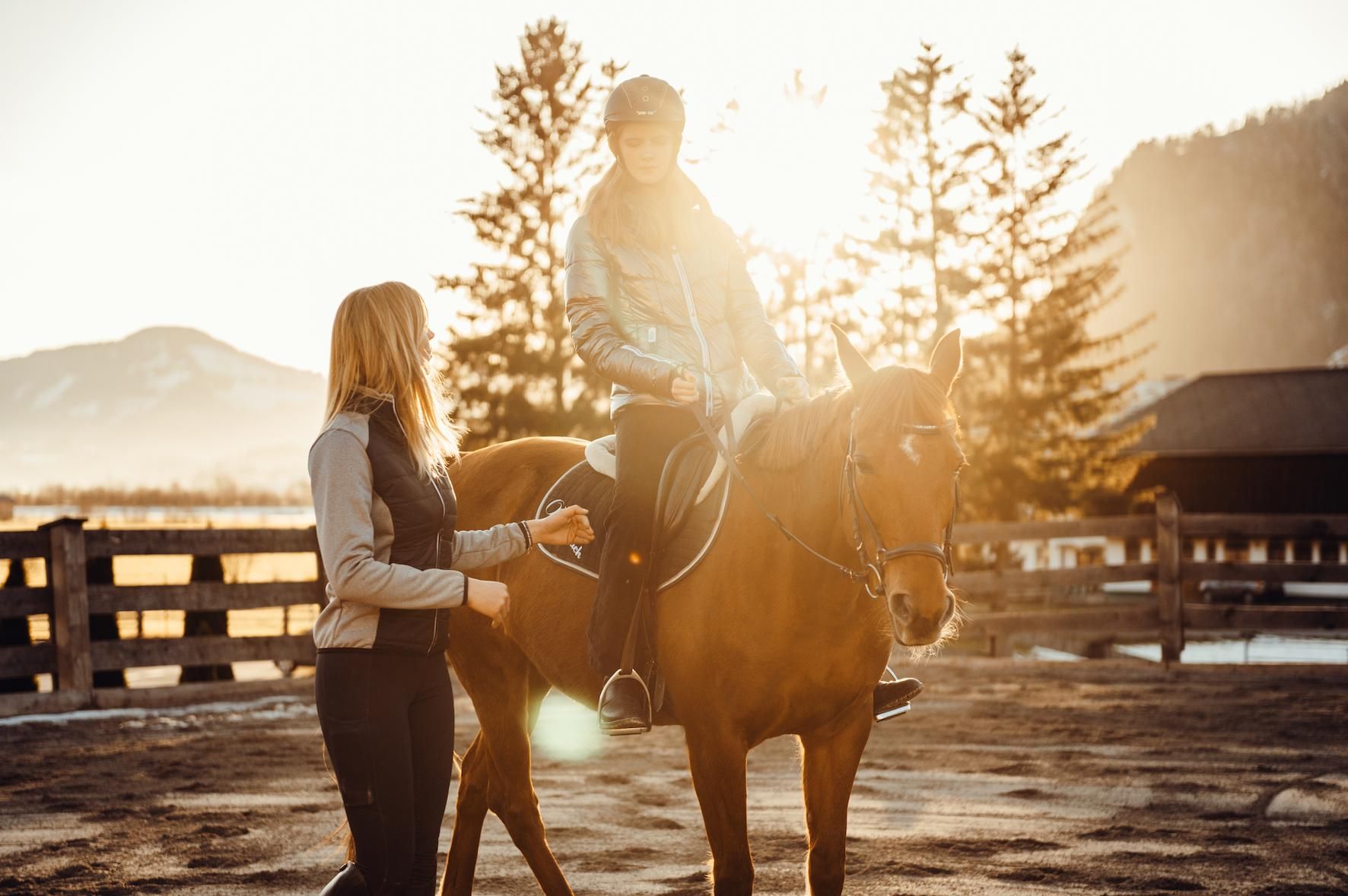 A woman is leading a horse on which a child is sitting. In the background, trees and gentle hills are visible at sunset.