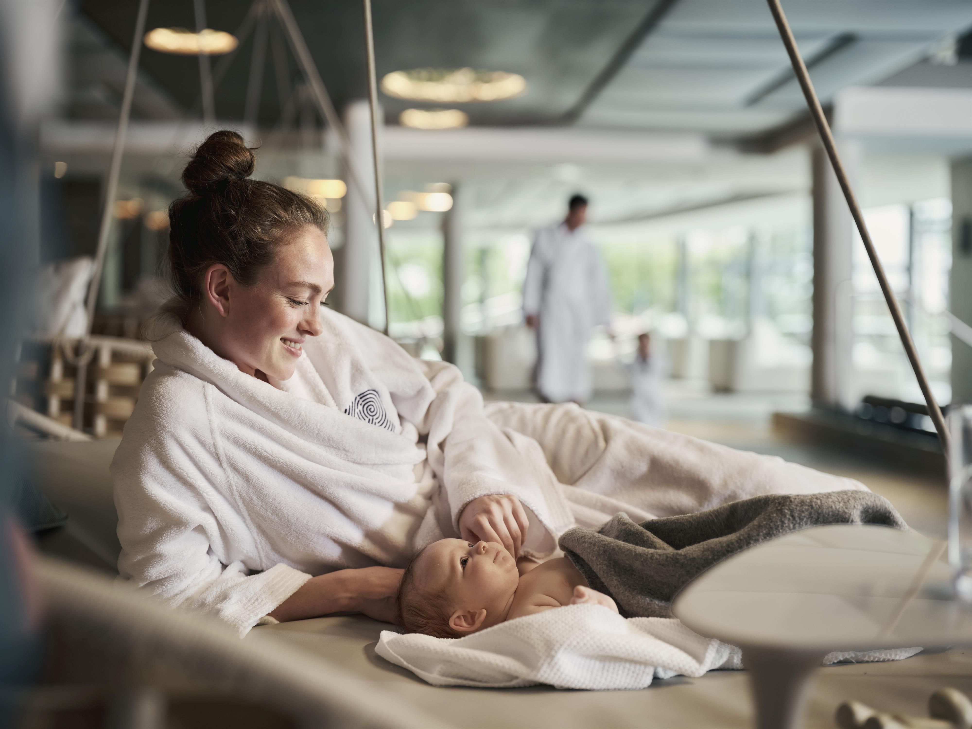 A young mother sits in a relaxing room, holding her baby in her arms. Both are wearing soft bathrobes and enjoying a quiet time together.