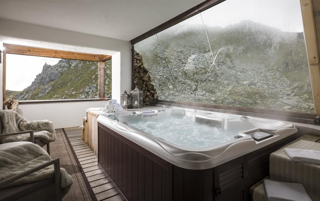 A hot tub in a modern room with large windows offers a view of a rocky mountain landscape. The room is comfortably designed with cozy furniture and a warm ambiance.