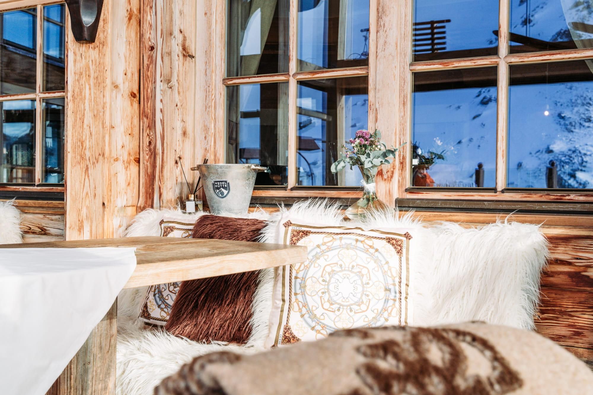 A cozy interior with wooden walls and large windows. There is a table with cushions and a small flower vase.