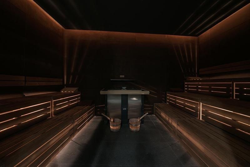 A modern sauna with dark wood and soft lighting. There are wooden benches and a central area with a heating source.
